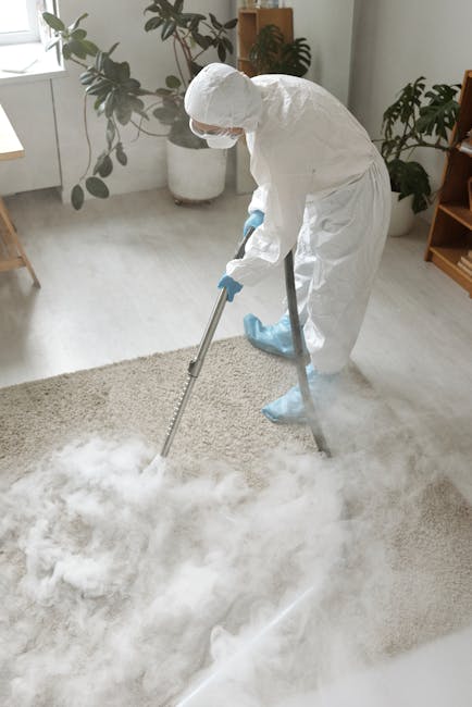 A person dressed in full protective cleaning gear, including a white coverall, gloves, mask, and shoe covers, is performing deep cleaning or sanitisation of a beige carpet in a bright, modern living r