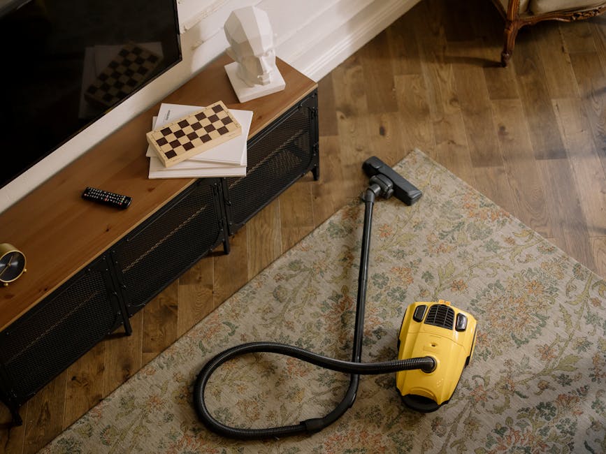 A person dressed in full protective cleaning gear, including a white coverall, gloves, mask, and shoe covers, is performing deep cleaning or sanitisation of a beige carpet in a bright, modern living r