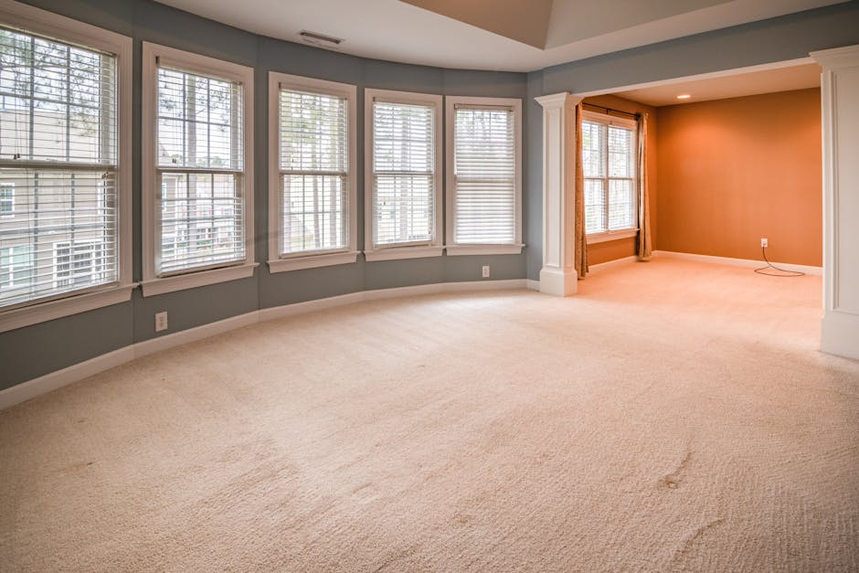 A person with curly dark hair wearing a green cleaning uniform is adjusting or preparing a textured, woven carpet runner in a living room. The room features a light grey upholstered sofa on wooden leg